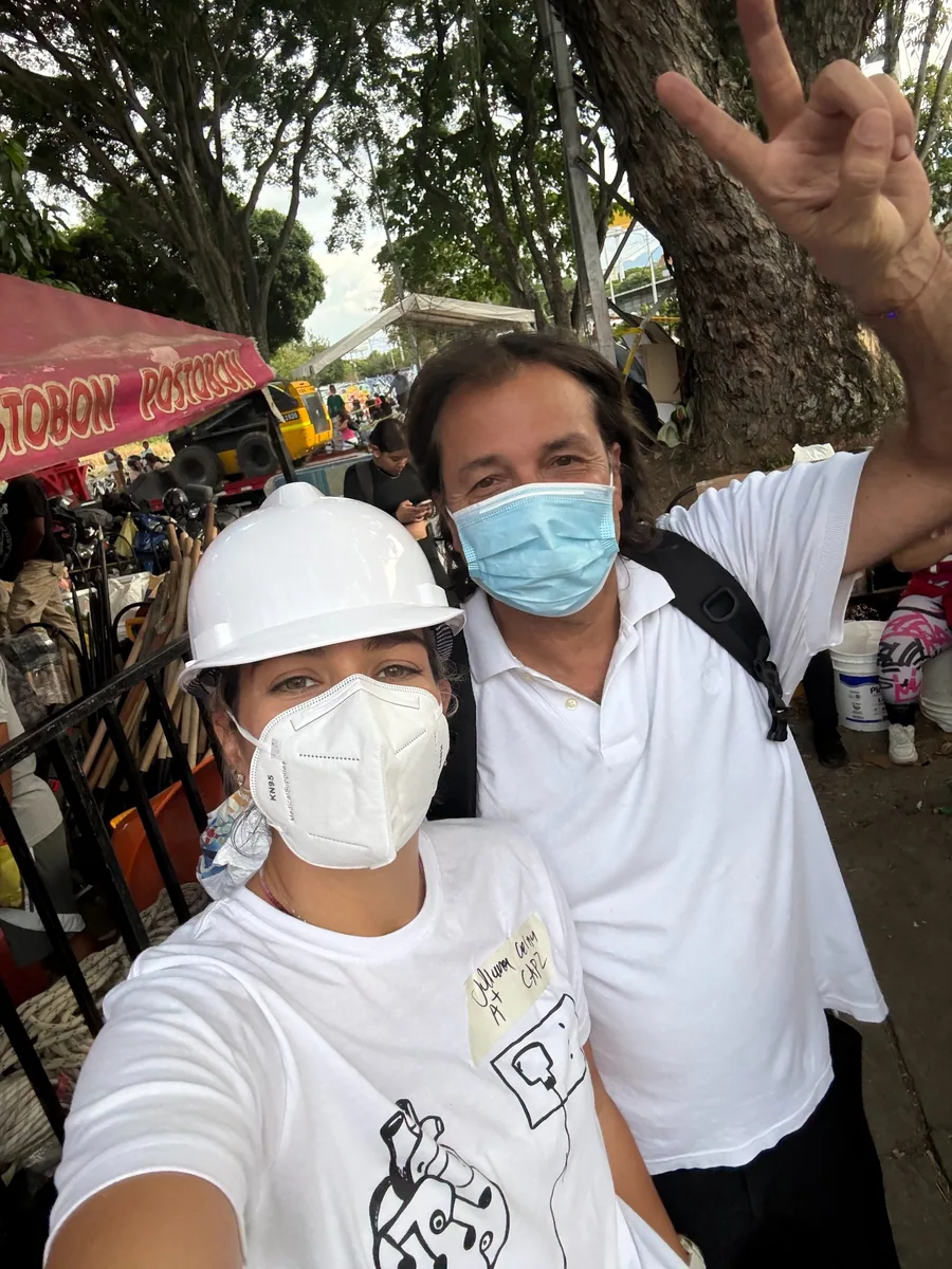 Dos personas con camisetas blancas y tapabocas, una de ellas con casco, al aire libre.