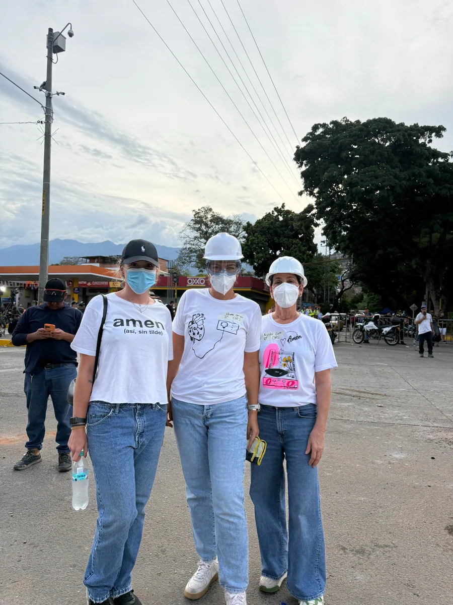 Tres personas con camisetas de AMEN, tapabocas y elementos de protección en una calle.
