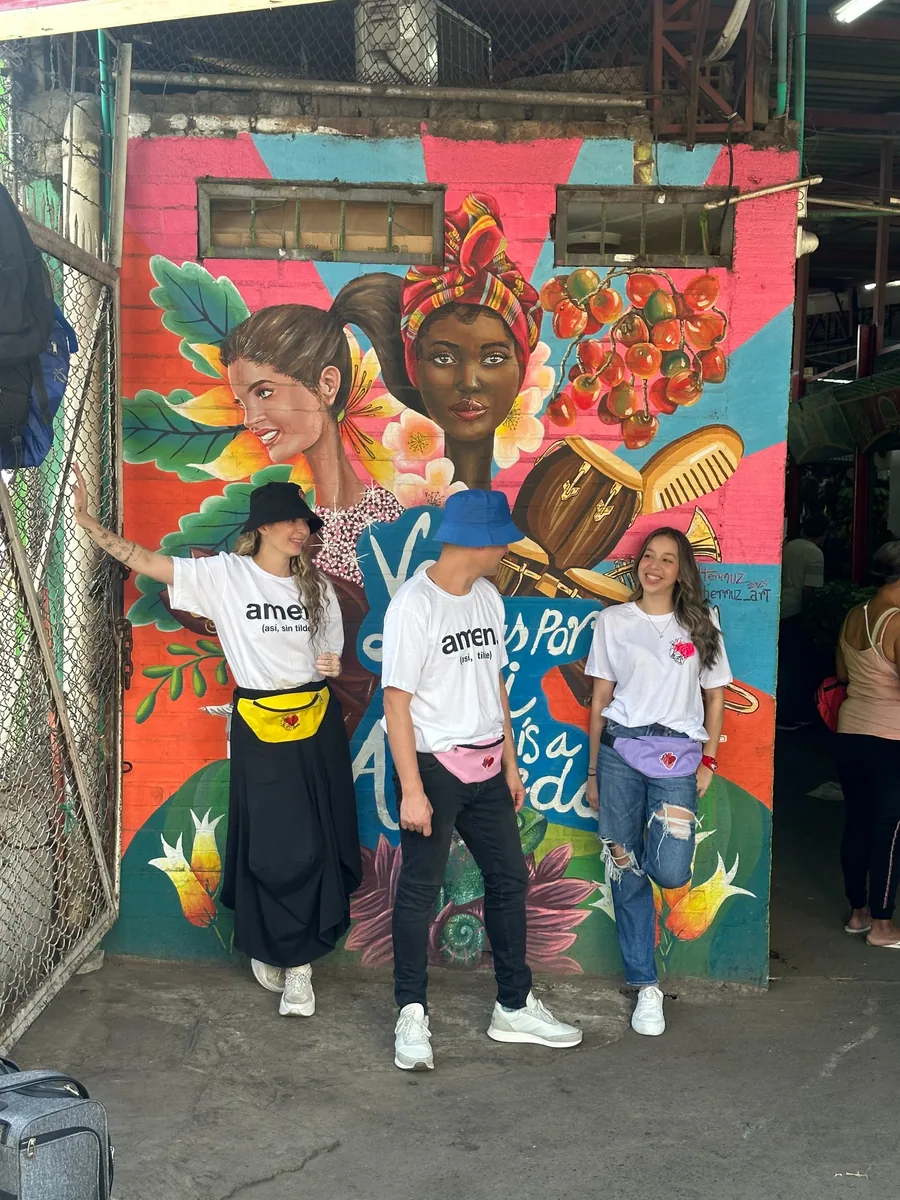 Tres personas con camisetas, canguros y sombreros de AMEN frente a un mural colorido.